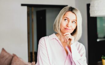 A woman with blonde hair and a thoughtful expression stands in a room. She is wearing a light pink blouse and has her hand on her chin. In the background, there is a sofa and a doorway.