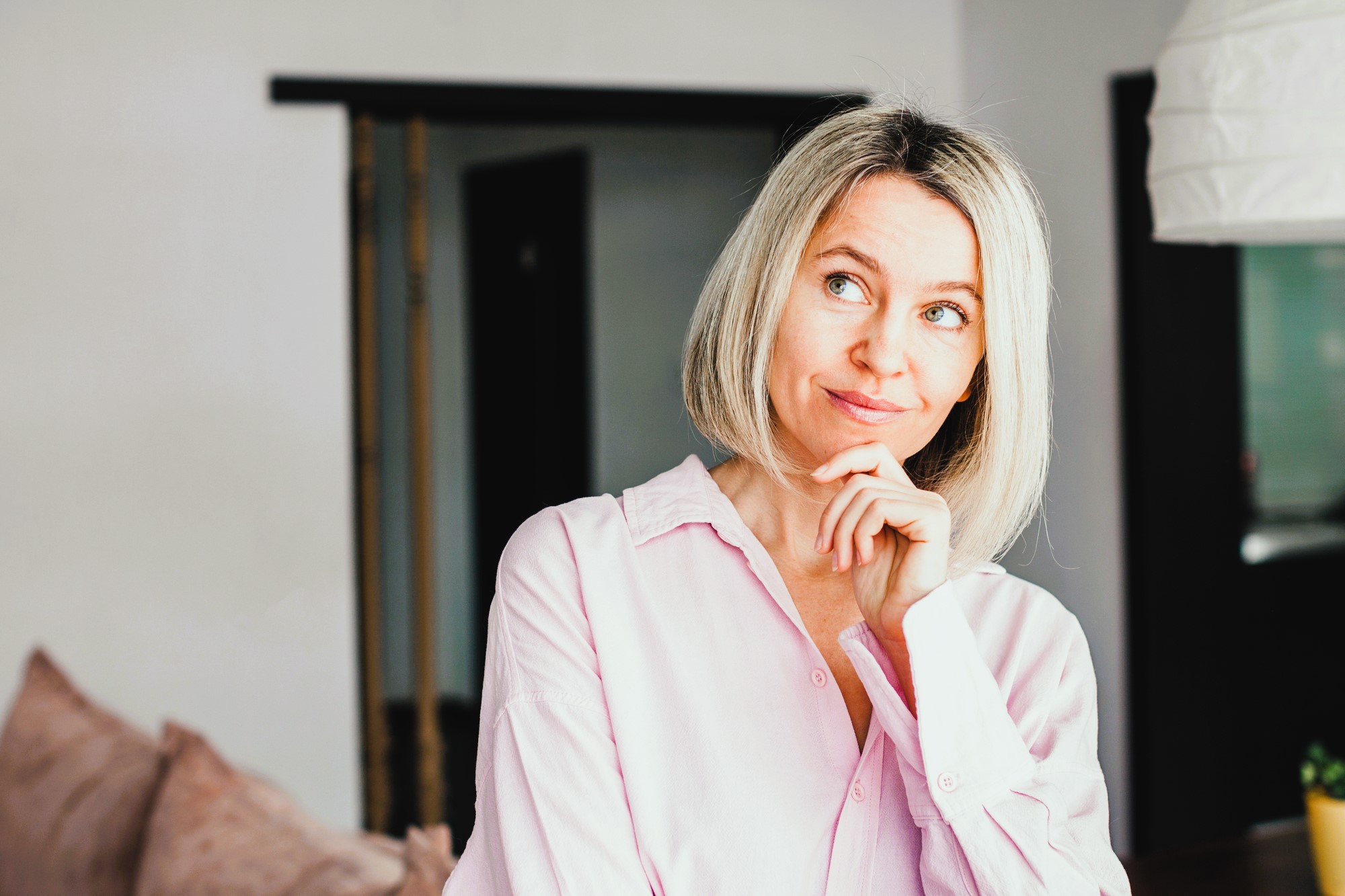 A woman with blonde hair and a thoughtful expression stands in a room. She is wearing a light pink blouse and has her hand on her chin. In the background, there is a sofa and a doorway.