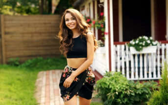 A smiling woman with long, wavy brown hair stands outside a house with a porch. She is wearing a black crop top and a floral skirt. The background shows a grassy yard, a wooden fence, and part of the house with flowers on the porch.