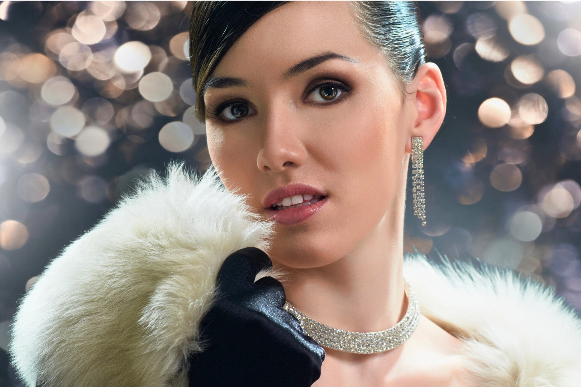 A woman with dark hair pulled back gazes at the camera. She wears a sparkling choker, long dangling earrings, and a white fur collar. The background is blurred with bokeh lights, creating a glamorous and elegant atmosphere.