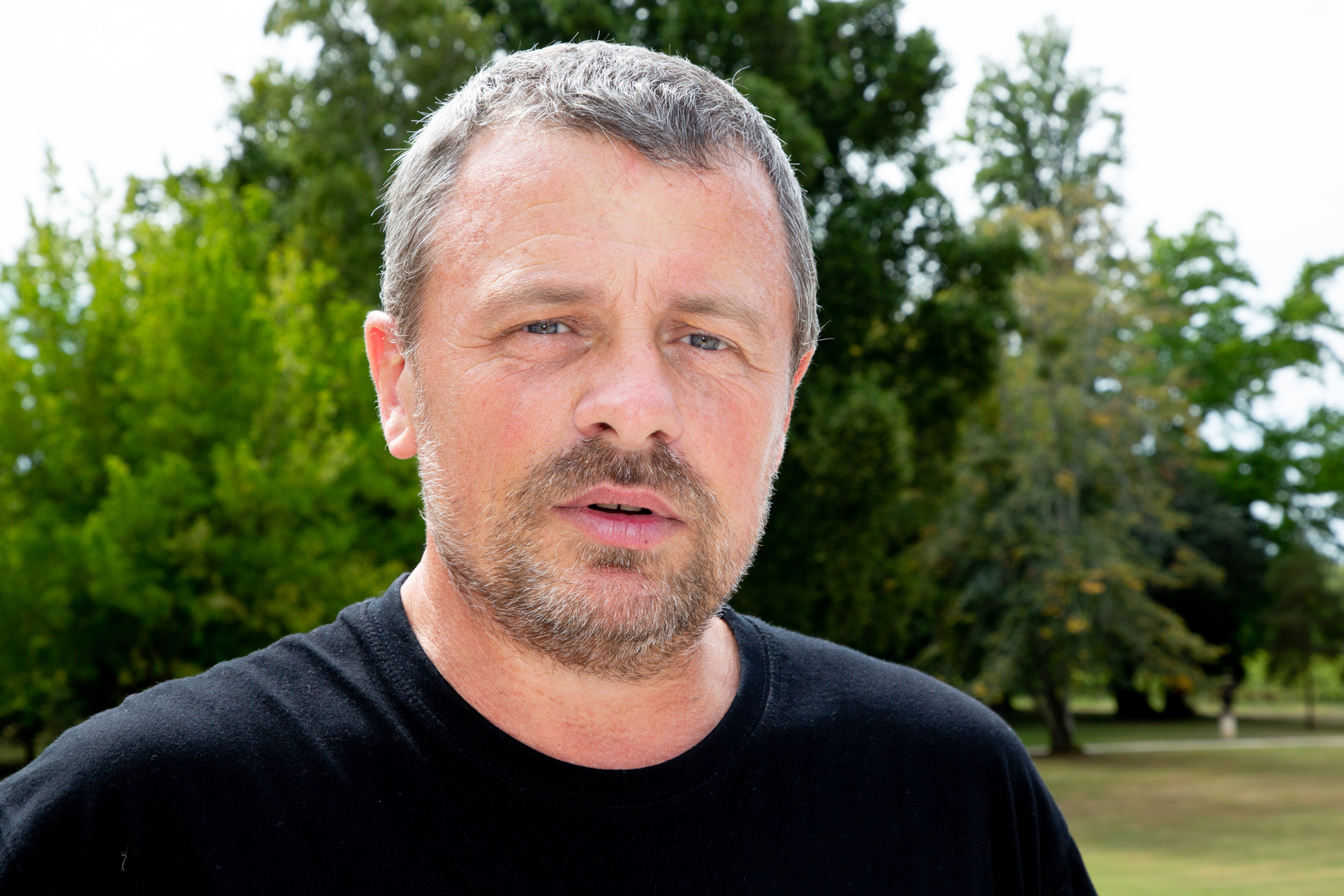 A man with short gray hair and a beard is standing outdoors. He is wearing a black t-shirt and looking directly at the camera with a neutral expression. The background features lush green trees and a bright sky.