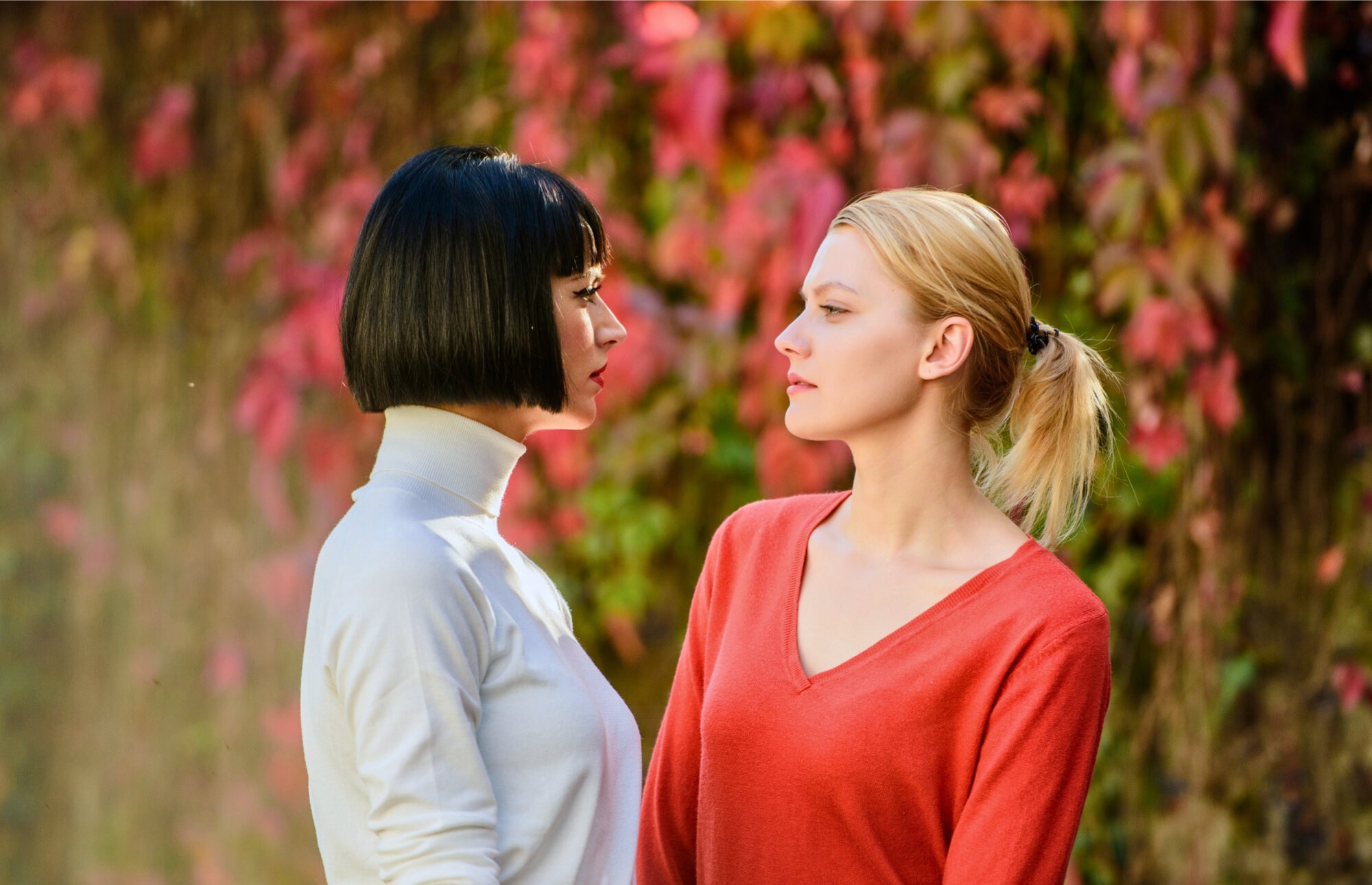 Two women stand outdoors, facing each other against a backdrop of vibrant autumn leaves. One has black hair and wears a white turtleneck, while the other has blonde hair tied back in a ponytail and wears a red V-neck top. Both have serious expressions.