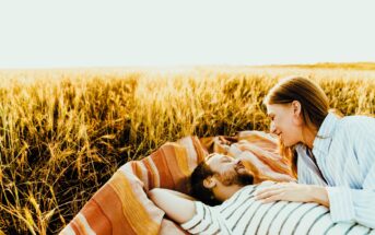 A couple relaxes on a striped blanket in a golden field. The man lies on his back with his arms behind his head, while the woman leans over him, smiling. Both are enjoying the sunny, serene atmosphere of the open field.