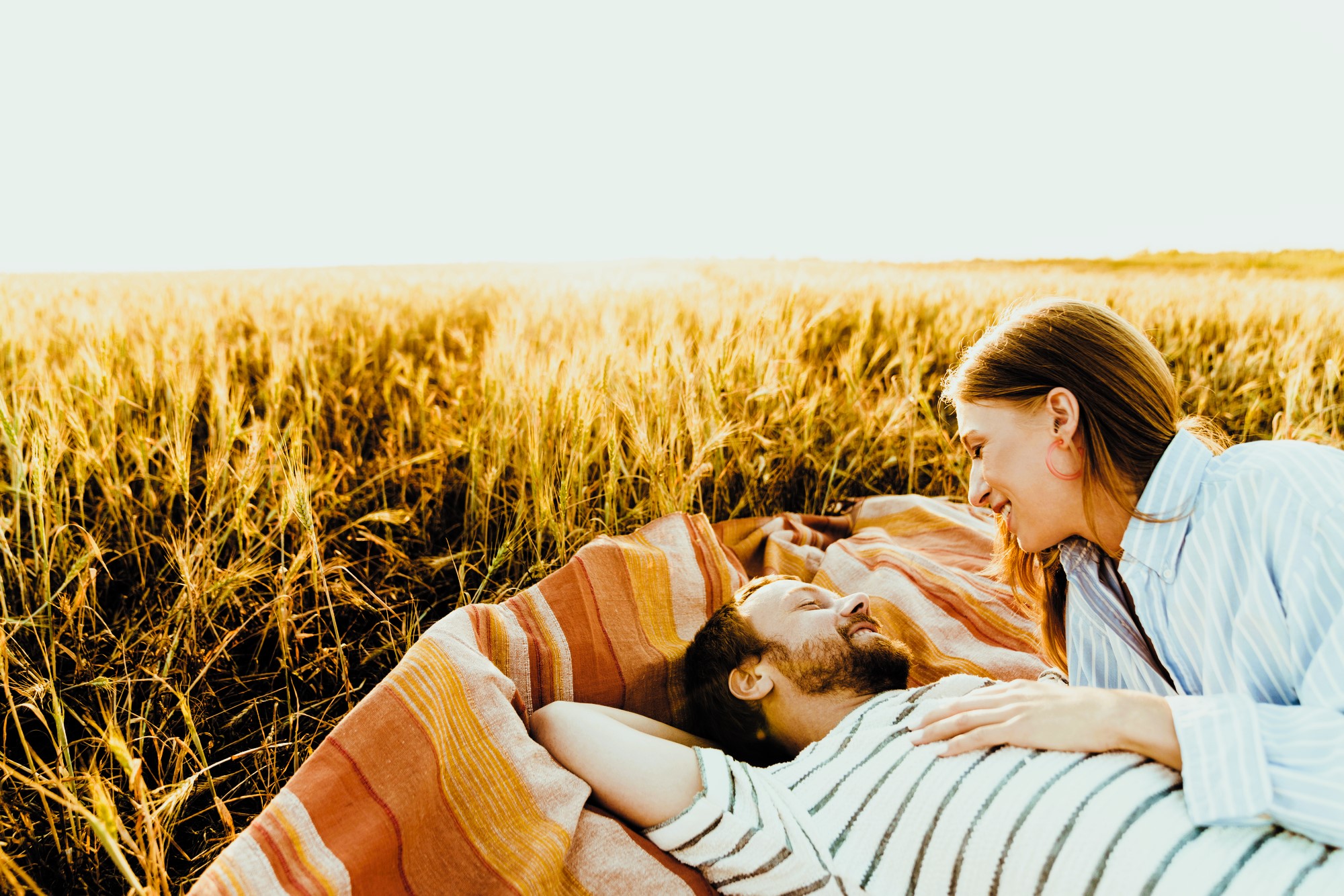 A couple relaxes on a striped blanket in a golden field. The man lies on his back with his arms behind his head, while the woman leans over him, smiling. Both are enjoying the sunny, serene atmosphere of the open field.