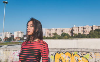 A woman with long dark hair stands outdoors against a backdrop of graffiti-covered walls and apartment buildings. She's wearing a red and black striped top and has her eyes closed, basking in the sunlight with a clear blue sky above.