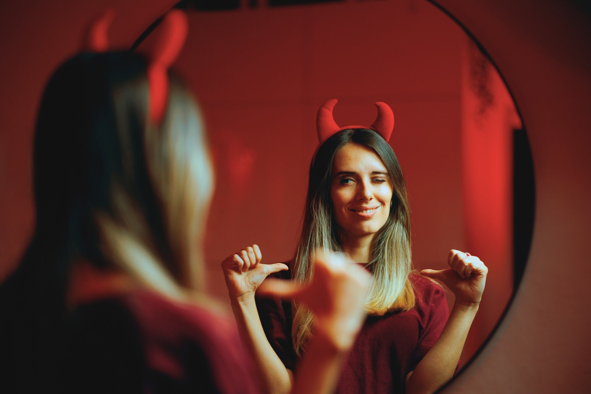 A woman with long hair, wearing red devil horns, stands in front of a round mirror. She is making a playful winking gesture and pointing at herself with both thumbs. The lighting in the room casts a red glow, adding to the playful atmosphere.