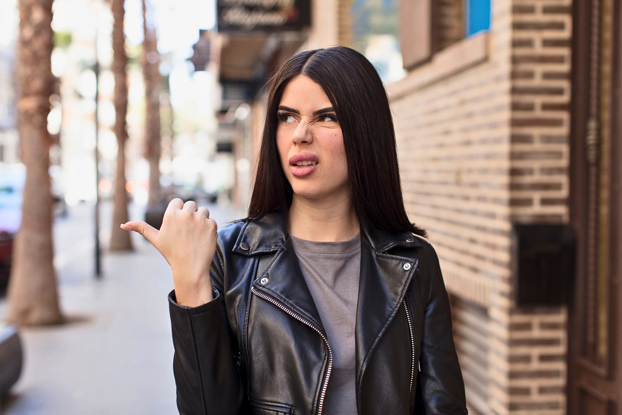 A woman with long dark hair, wearing a black leather jacket, is standing on an outdoor urban street lined with brick buildings and palm trees. She is looking to her right with a puzzled expression, her left thumb pointing behind her.