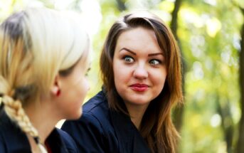 Two women are engaged in conversation outdoors. One woman, with dark hair and expressive eyes, has a surprised or amused look on her face. The other woman, with blonde hair tied in a braid, is facing the first woman and appears to be speaking. They are in a green, leafy setting.