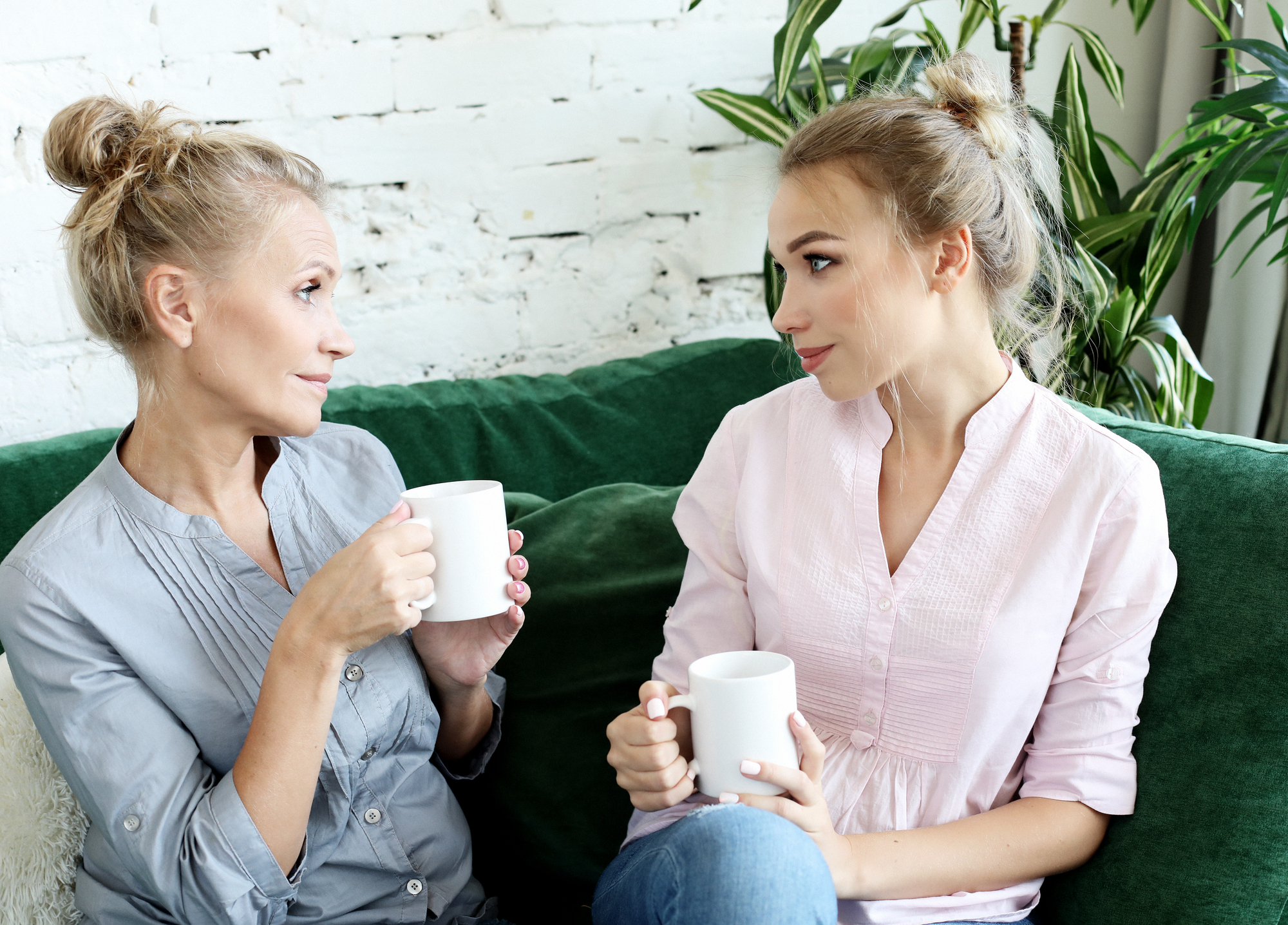 Two women sit on a green couch, engaged in conversation and holding white mugs. Both have blonde hair, styled in high buns. One wears a light grey blouse, and the other wears a pale pink blouse. A white brick wall and green plants are in the background.