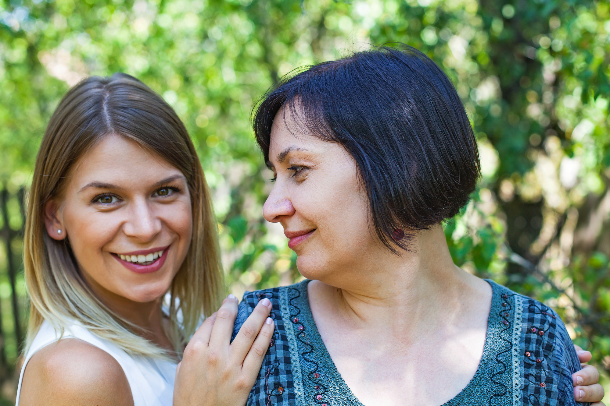 Two women are outdoors with green foliage in the background. The woman on the left has long, blonde hair and is smiling at the camera. The woman on the right has short, dark hair and is looking at the woman on the left with a gentle expression.