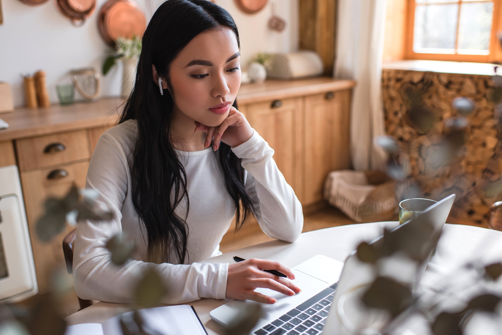 A young woman with long dark hair sits at a table in a cozy kitchen, wearing a white long-sleeve shirt and wireless earbuds. She is using a laptop, looking focused and thoughtful, with her chin resting on her hand. A window and wooden cabinets are in the background.