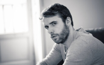 A black-and-white photo shows a man with short hair and a beard. He is wearing a light-colored shirt and is seated, leaning forward slightly, with a thoughtful expression on his face. The background is softly lit, with a window visible behind him.