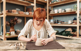 A person with short red hair is meticulously sculpting a clay pot on a table in a pottery studio. The individual is dressed in a white, long-sleeved shirt and surrounded by various pottery tools and shelves filled with pottery items in the background.