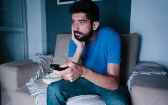 A man with a beard and mustache sits on a couch in a dimly lit room, holding a TV remote in one hand and resting his head on the other. He is wearing a blue t-shirt and jeans. A bowl of popcorn is on the couch beside him. His expression appears bored or disinterested.