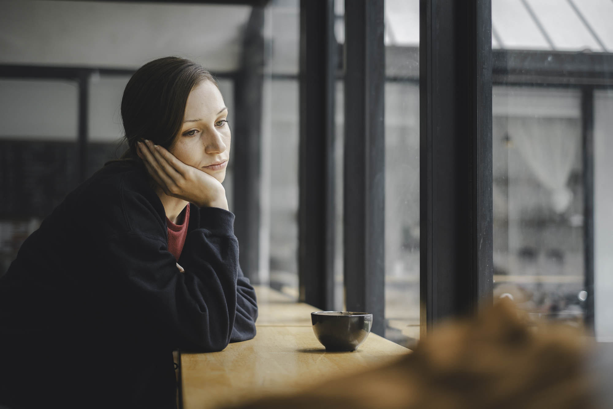 A woman with long brown hair rests her head on her hand, gazing out of a large window. She appears thoughtful or reflective. She is seated at a wooden countertop in what seems to be a café, with a small, dark cup in front of her. Natural light illuminates the scene.