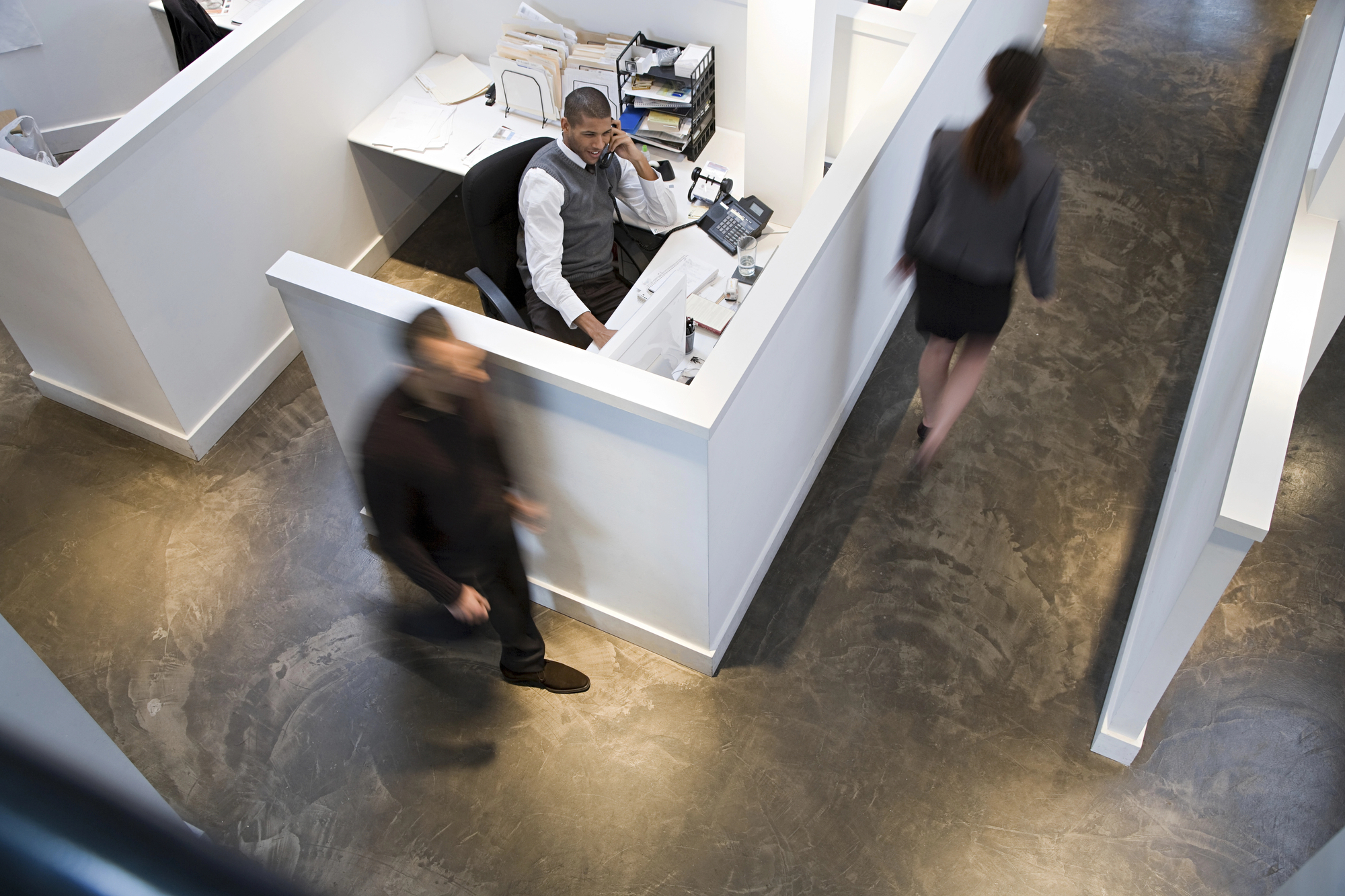 An office scene with a person sitting at a desk in a cubicle, speaking on the phone and looking at a computer monitor. Two people are walking past the cubicle in opposite directions. The floor is concrete, and papers and office supplies are scattered around the desk.