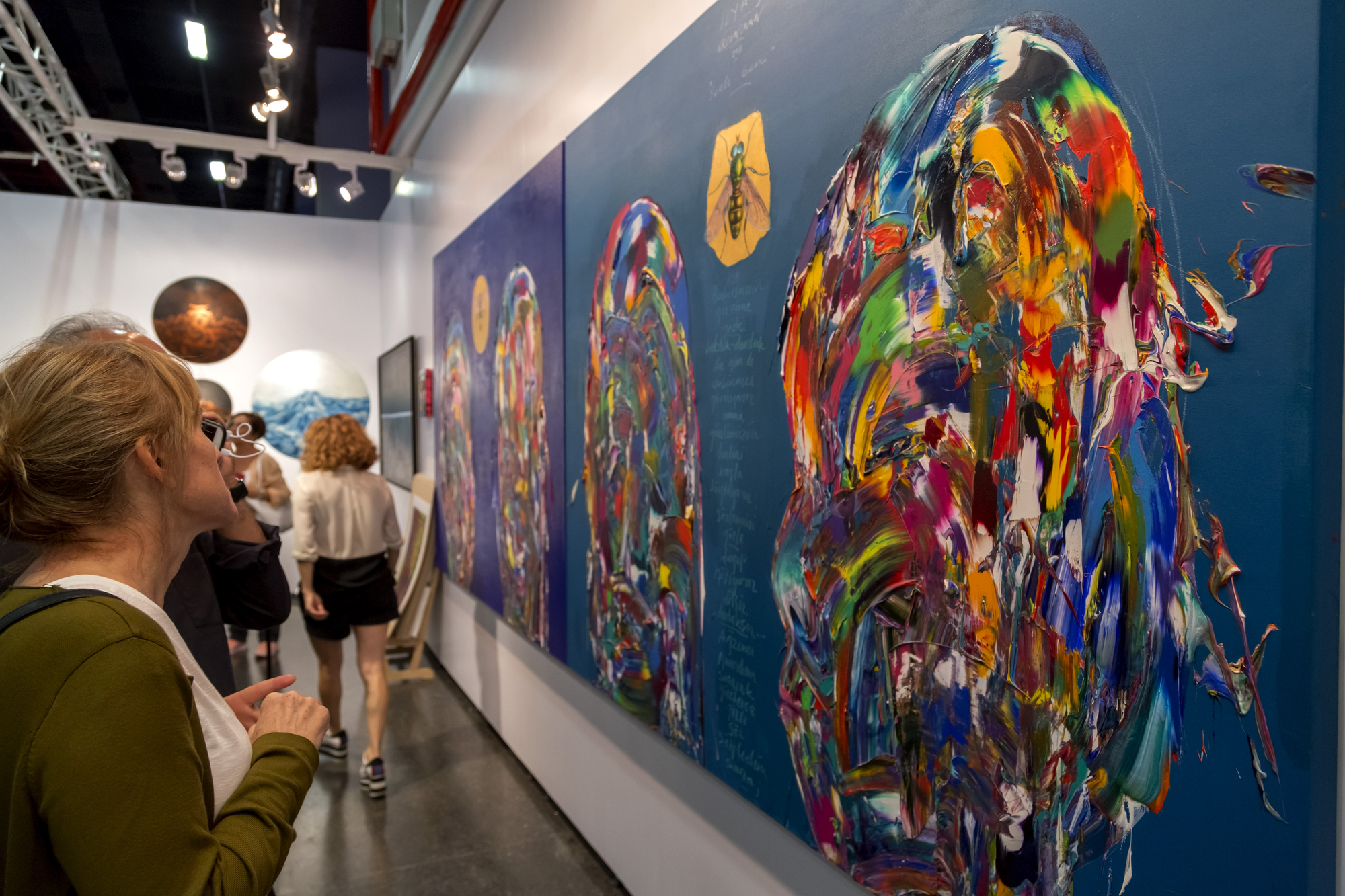 People observe colorful abstract paintings displayed on the walls of an art gallery. The paintings have vibrant swirls and splashes of color. The surroundings are modern with other visitors visible in the hallway, some engaging with the artwork.