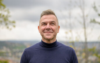 A man with short, gray hair and a slight smile is looking up. He is wearing a dark blue turtleneck sweater. In the blurred background, there are trees and a cloudy sky.