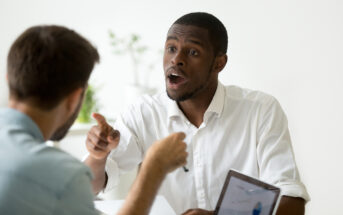 A man in a white shirt, appearing frustrated or upset, gestures with his hand and points while speaking to another man. The other man, seen from the back, is holding a pen. A laptop with a pie chart is also visible on the table between them.