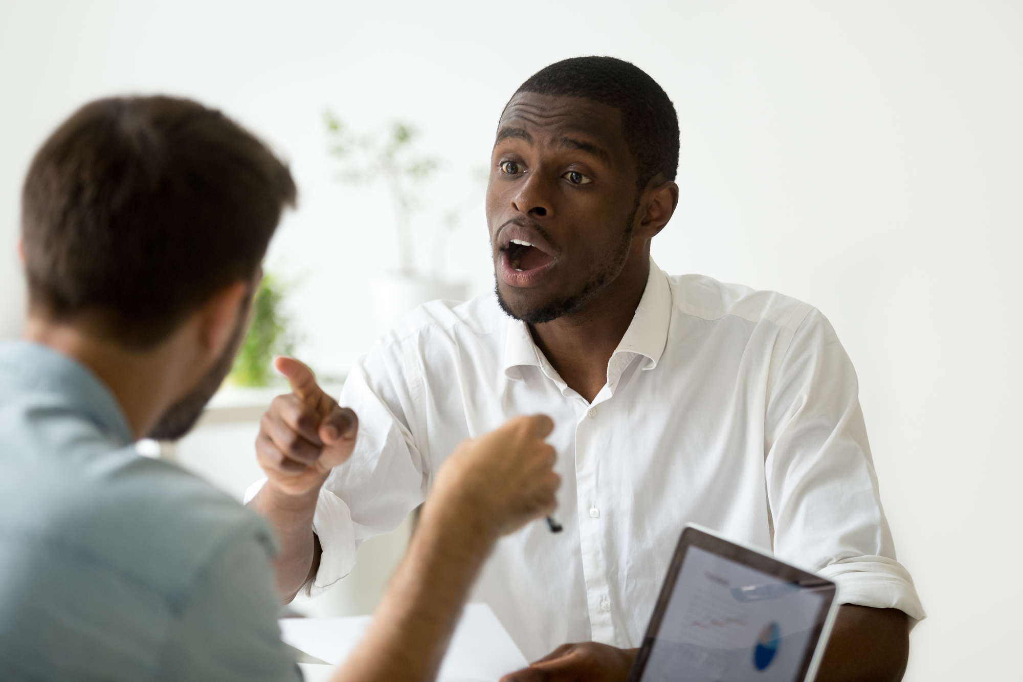 A man in a white shirt, appearing frustrated or upset, gestures with his hand and points while speaking to another man. The other man, seen from the back, is holding a pen. A laptop with a pie chart is also visible on the table between them.