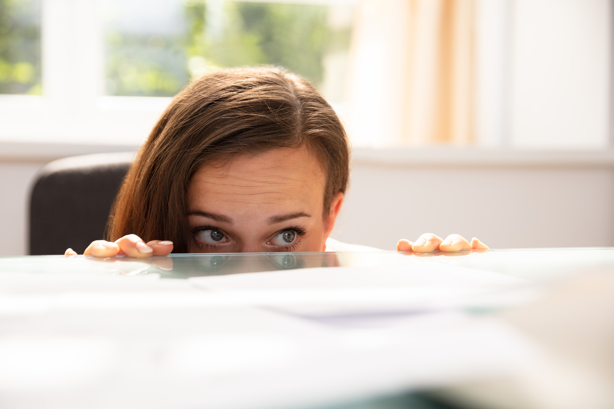 A woman with brown hair peeking over a table, her eyes wide open in a curious or cautious expression. She is partially hidden behind the table, with her hands gripping the edge. The background shows a window with blurred greenery outside.