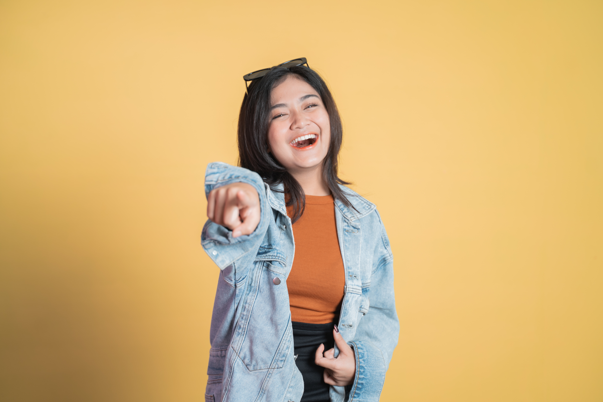 A young woman with long dark hair, wearing a denim jacket and an orange top, smiles happily while pointing towards the camera. She has a pair of sunglasses resting on her head. The background is a solid yellow color.