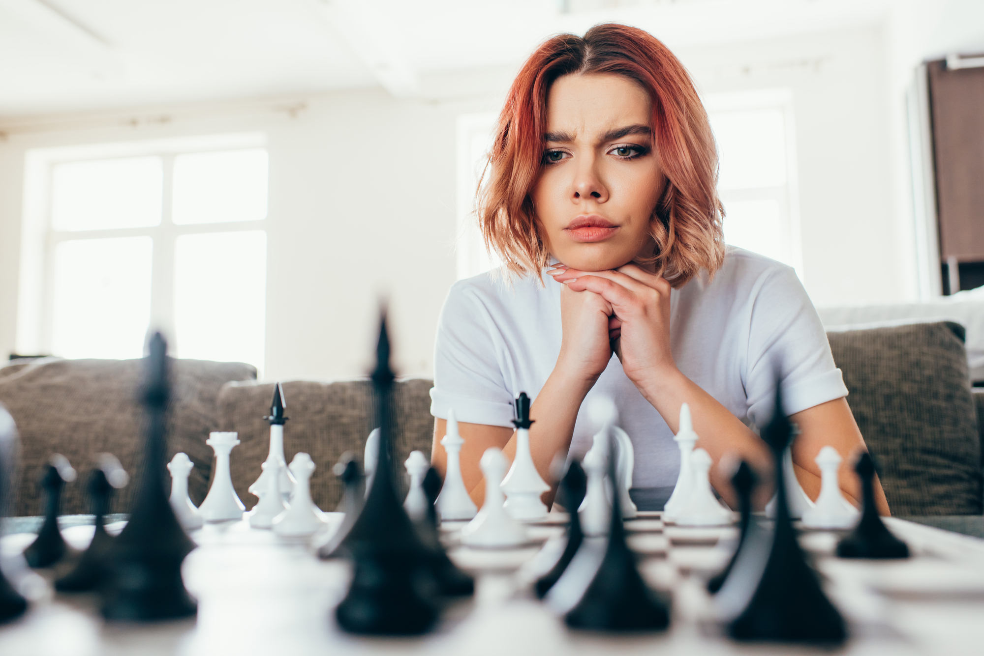 A woman with chin resting on her hands looks thoughtfully at a chessboard in front of her. She has shoulder-length reddish hair and wears a white shirt. The room is bright with sunlight streaming through large windows in the background.