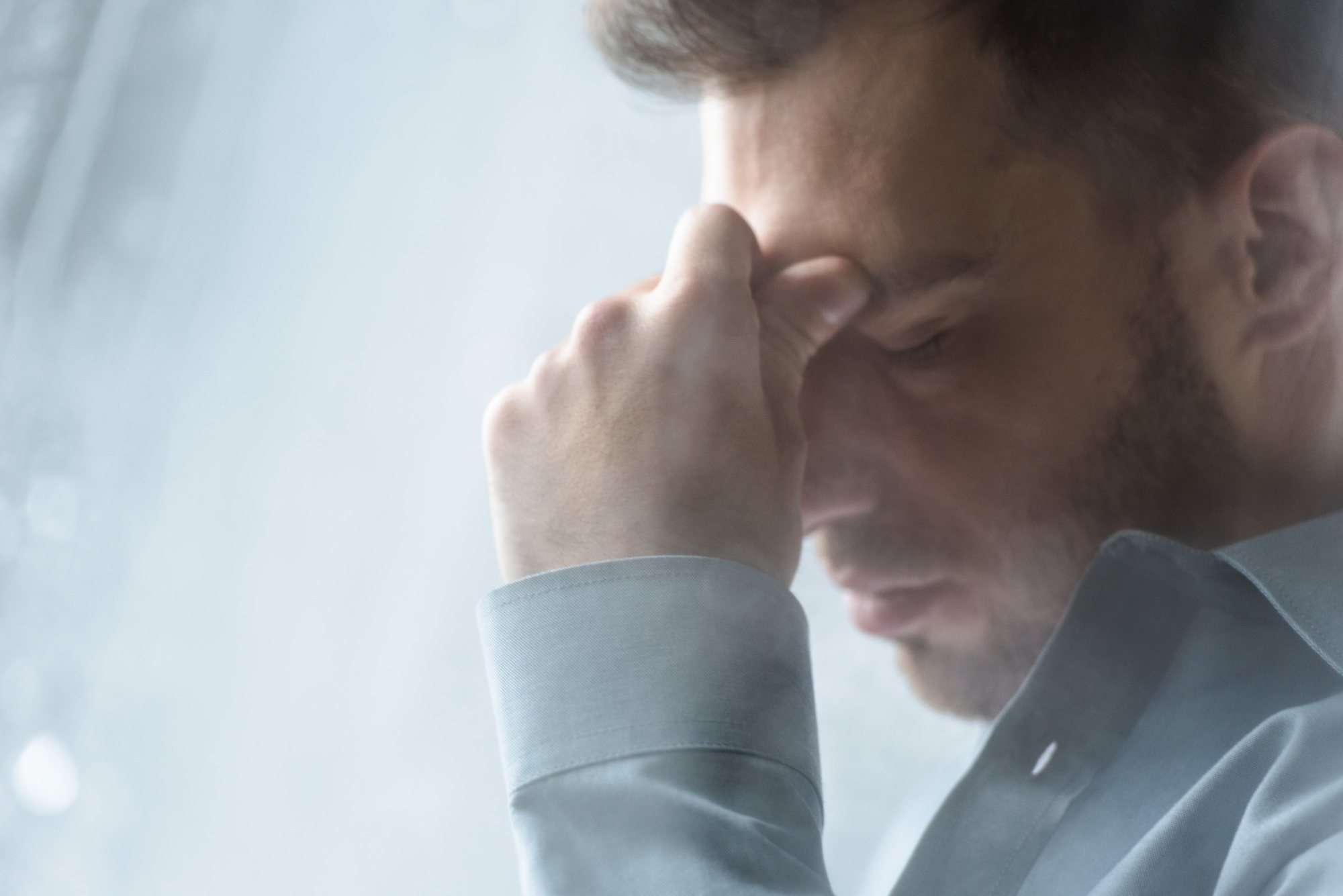 A man in a light blue shirt is shown from the shoulders up, with a blurred background. He has his eyes closed and his hand pressed against his forehead, appearing stressed or deep in thought. The image has soft lighting and a contemplative mood.