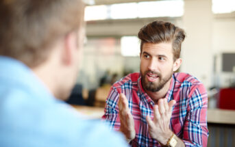 A bearded man in a plaid shirt gestures expressively with his hands while engaged in conversation with another person, who is partially visible and wearing a blue shirt. They are in a brightly lit indoor setting with blurred office elements in the background.