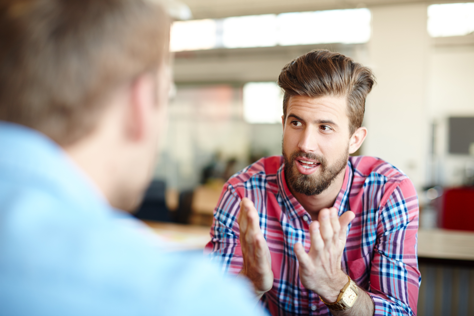 A bearded man in a plaid shirt gestures expressively with his hands while engaged in conversation with another person, who is partially visible and wearing a blue shirt. They are in a brightly lit indoor setting with blurred office elements in the background.