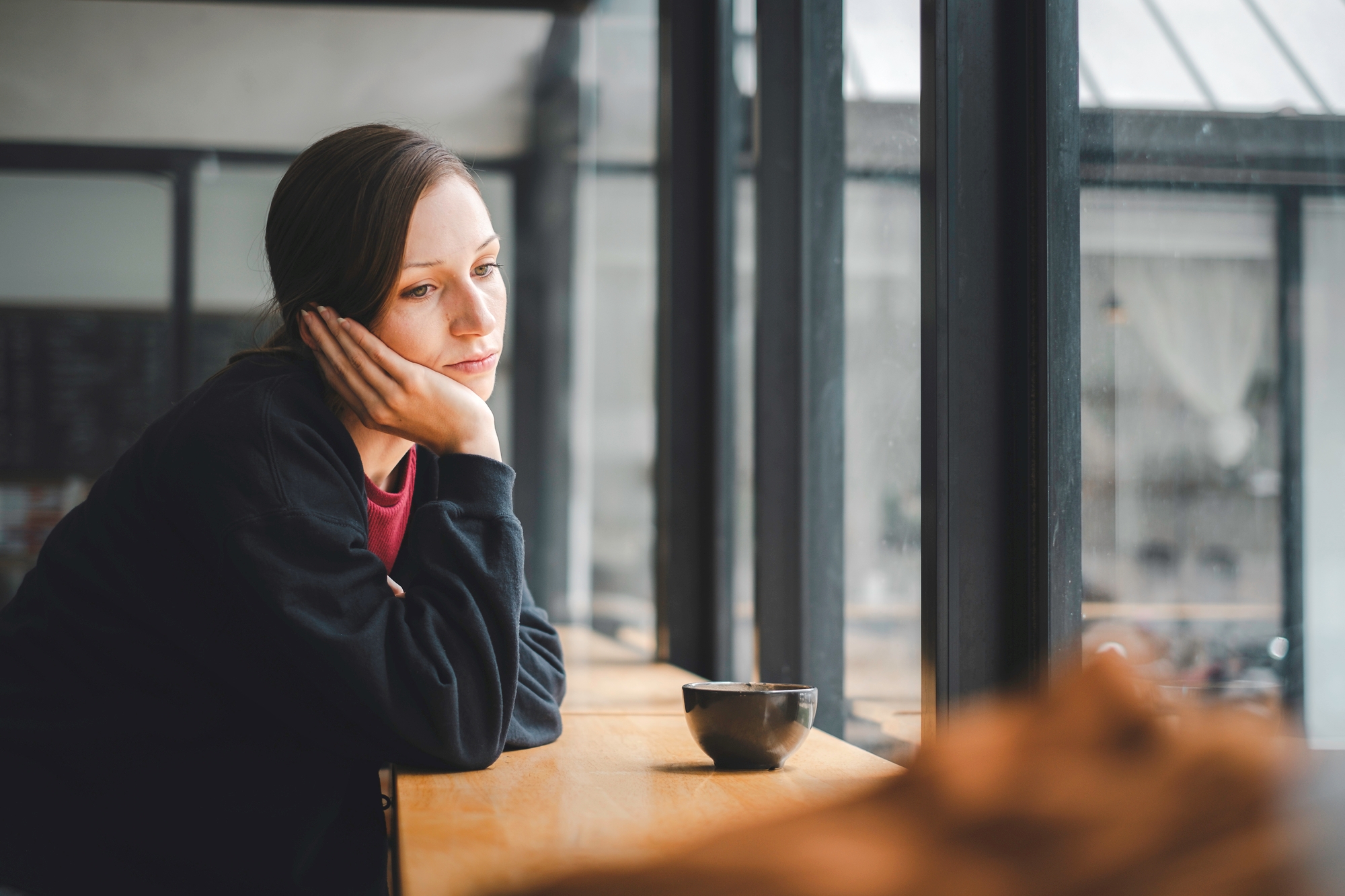 A woman sits alone at a wooden countertop, resting her chin on her hand and gazing out a large window with a pensive expression. She has a bowl next to her, and soft daylight filters through the window. The setting appears to be a cozy café or coffee shop.