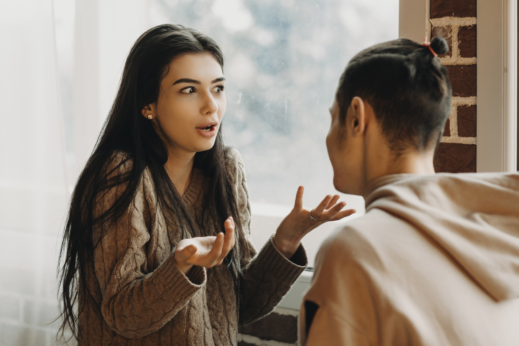 A woman with long dark hair, wearing a brown sweater, gestures with her hands while speaking to a person with short hair and a topknot, who is facing away from the camera. They are in a brightly lit room with a brick wall and window in the background.