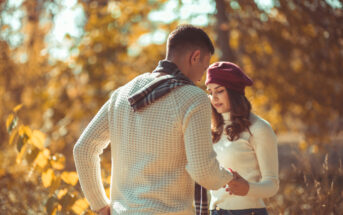 A couple stands outdoors among autumn foliage. The man, in a white sweater and scarf, gently holds the hand of the woman, who wears a white sweater and a red beret. They're engaged in a quiet, intimate moment with warm sunlight filtering through the trees.