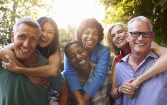 A diverse group of six adults smiling and posing cheerfully outdoors, with sunlight streaming through trees in the background. Two individuals in the front, one man, and one woman are being hugged from behind by two others. The atmosphere is lively and joyful.