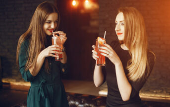 Two women are standing inside a dimly-lit space with exposed brick walls, each holding and sipping colorful cocktails. They are smiling and appear to be engaged in conversation, creating a warm and relaxed atmosphere.