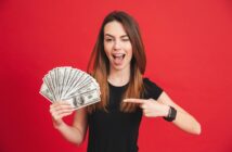 A woman with long brown hair wearing a black t-shirt and a smartwatch is standing in front of a red background. She is holding a fanned-out stack of 100-dollar bills in one hand and pointing to it with the other. She is winking and smiling.