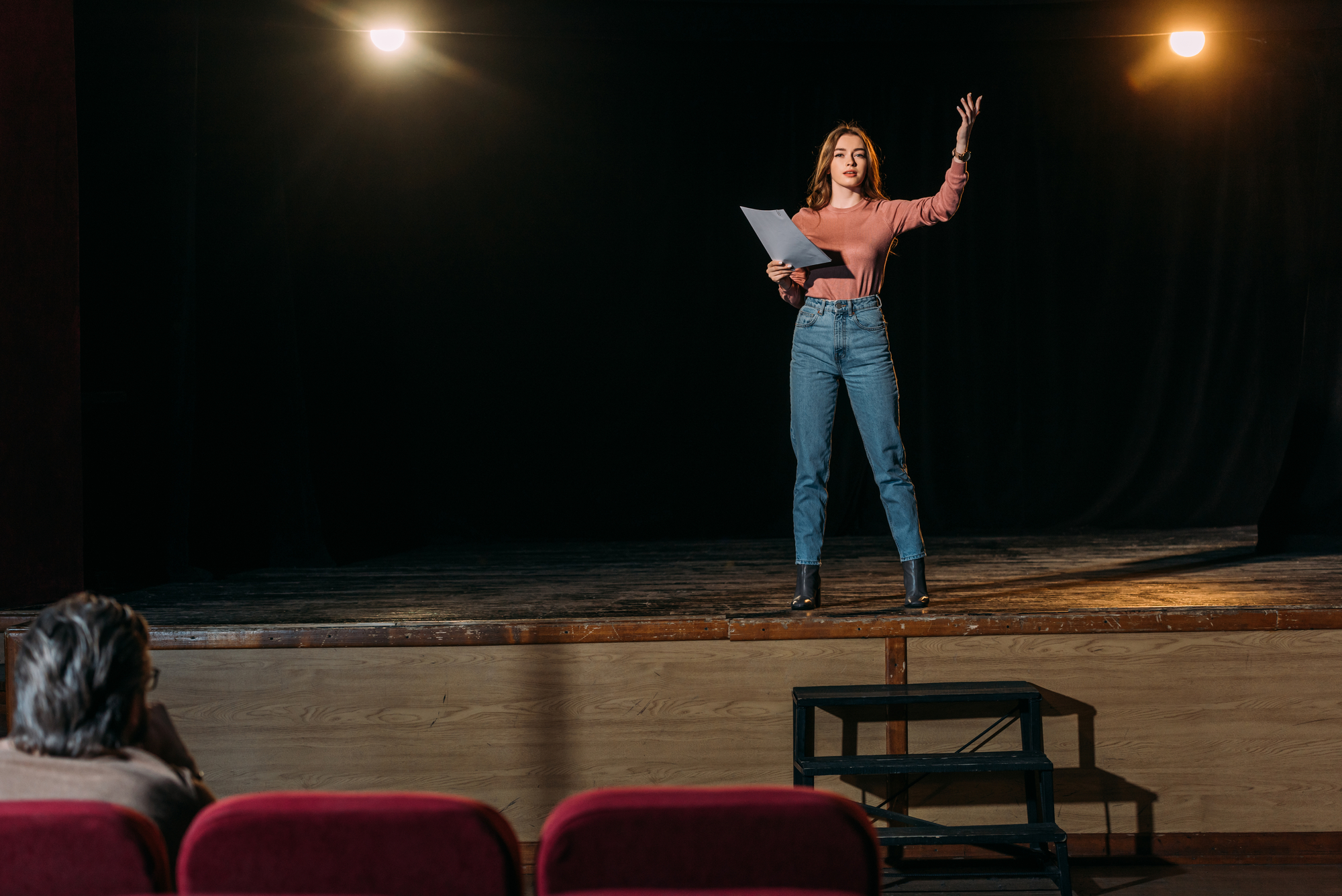 A woman on a dimly lit stage performs while holding a script in one hand and gesturing with the other. She wears a long-sleeve shirt and jeans. In the foreground, a person watches from the audience. Red theater seats and stage lights are visible.