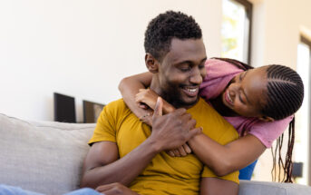 A man in a yellow shirt sits on a couch, smiling warmly as a young girl in a pink shirt hugs him from behind, both appearing joyful and affectionate. The background shows a bright, modern living room with large windows.