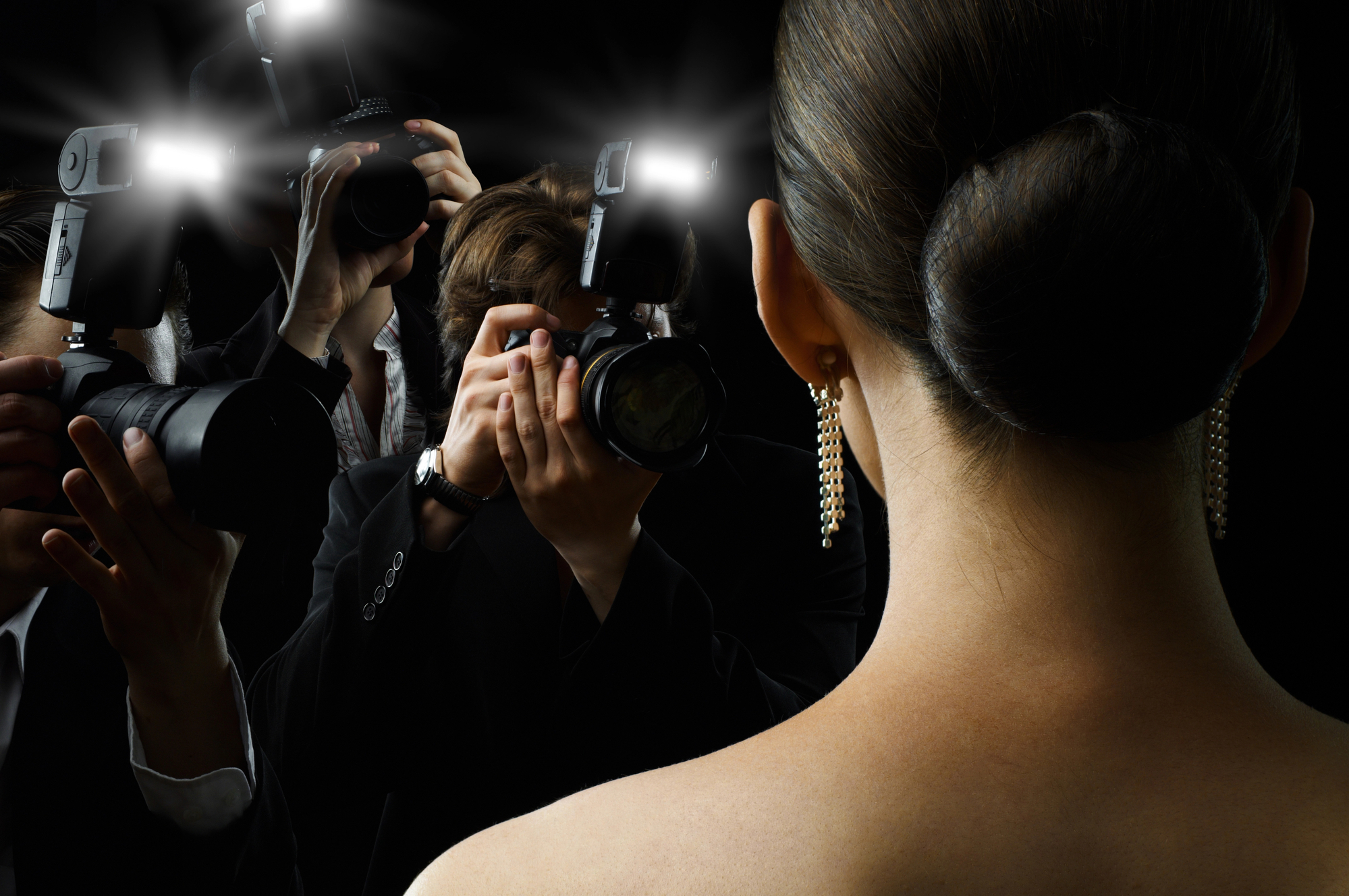 A person with their dark hair tied in a bun is facing away from the camera, wearing dangly earrings. They are surrounded by photographers taking pictures, with bright camera flashes illuminating the scene against a dark background.