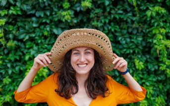 A smiling woman stands in front of a lush green background, wearing a large woven straw hat and an orange top. She has long, wavy brown hair and is holding the brim of the hat with both hands.