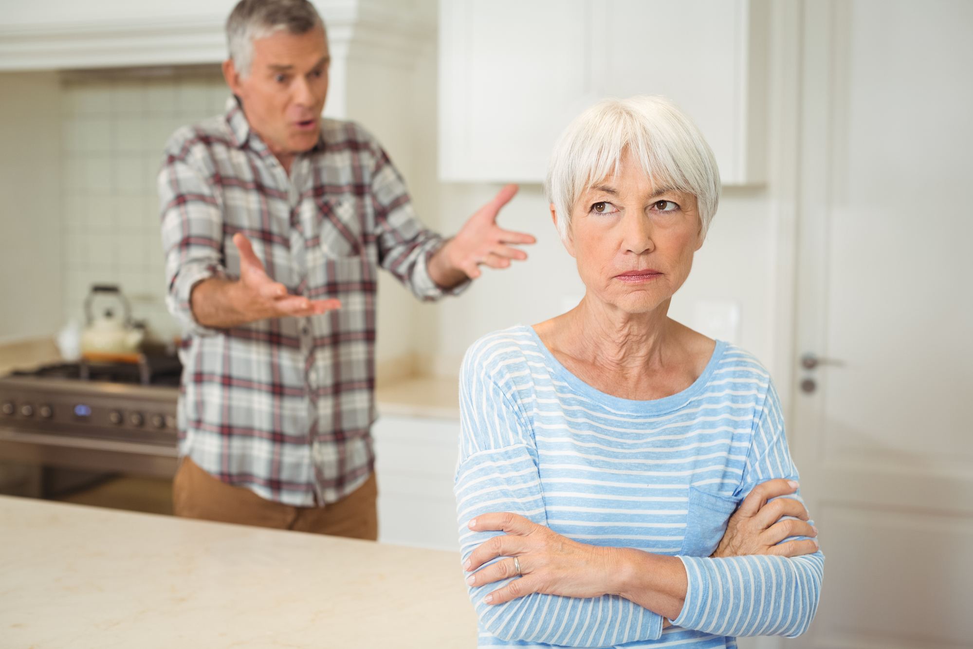 An older woman with short white hair and a blue striped shirt stands with her arms crossed, looking upset. In the background, an older man with gray hair and a plaid shirt gestures animatedly, appearing to speak earnestly or argue. They are in a kitchen.