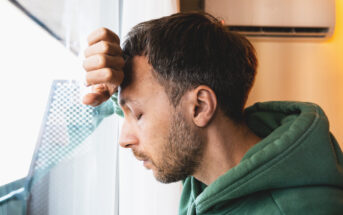 A person wearing a green hoodie leans their forehead against a glass window, eyes closed and with a pensive expression. The background shows an air conditioning unit and part of a balcony or exterior structure.