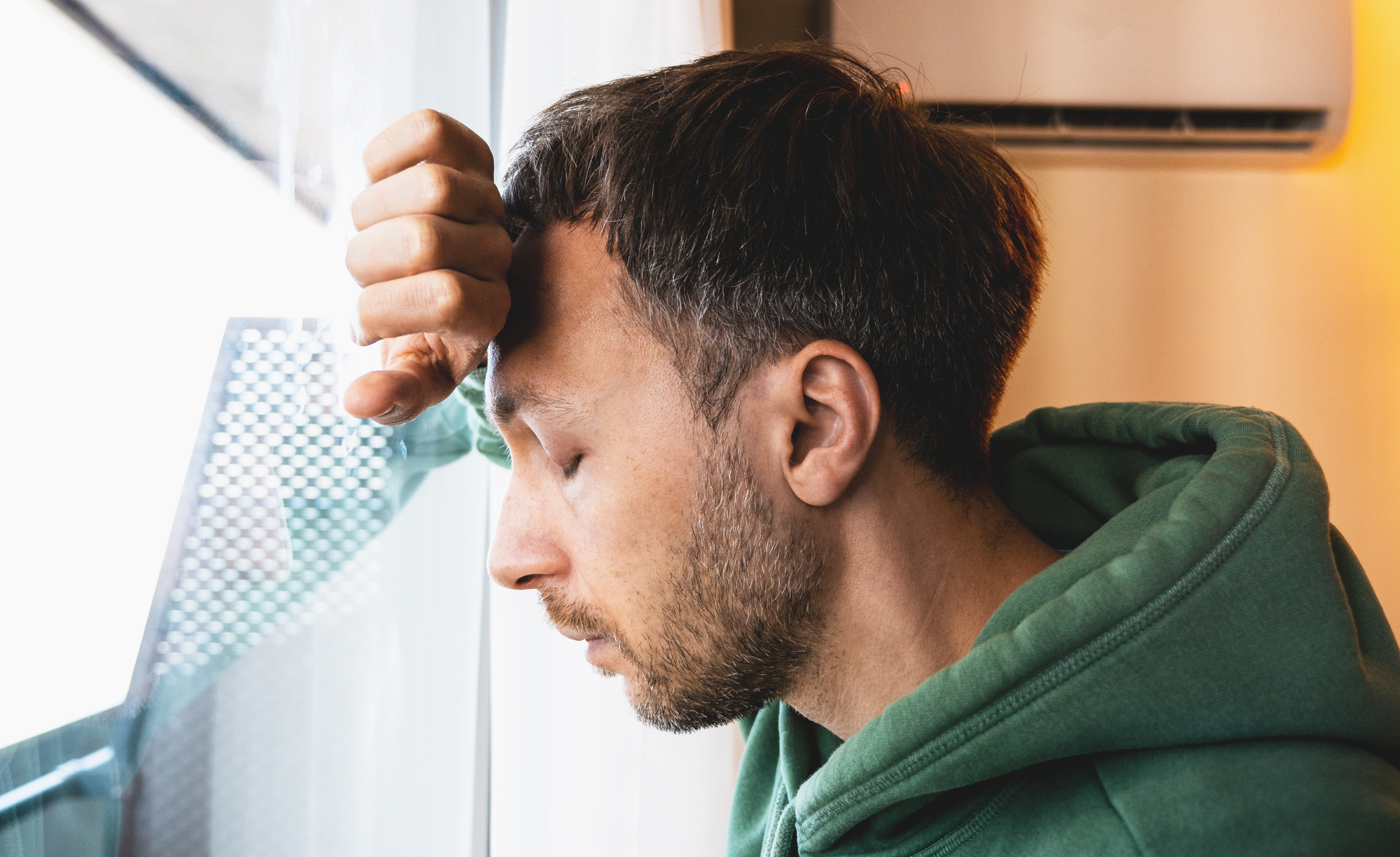 A person wearing a green hoodie leans their forehead against a glass window, eyes closed and with a pensive expression. The background shows an air conditioning unit and part of a balcony or exterior structure.