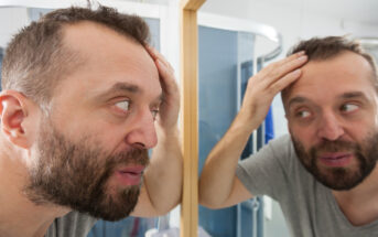 A man with a beard and short hair examines his reflection closely in a bathroom mirror, looking concerned. He is holding his head with both hands, focusing on the top of his head. The bathroom features a modern shower enclosure in the background.