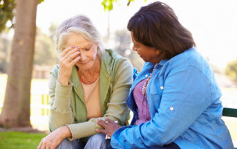 An elderly woman in a light green hoodie sits on a bench outdoors, resting her face in her hand, while a middle-aged woman in a blue jacket sits beside her, offering comfort by gently placing her hand on the elderly woman's arm. They appear to be in a park.