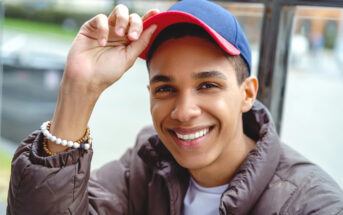 A smiling person wearing a blue and red baseball cap and a brown jacket is adjusting their cap with one hand. They have a beaded bracelet on their wrist and are sitting outdoors.