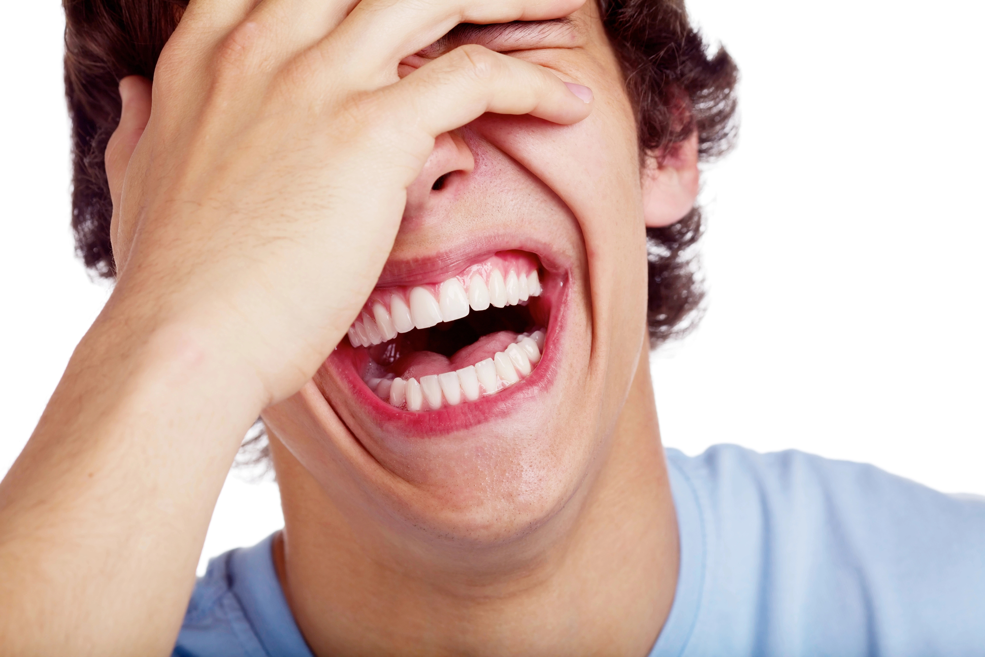 A person with dark hair is laughing heartily, with their mouth open wide, displaying their teeth. They are covering most of their face with one hand, and their eyes are closed. They are wearing a light blue shirt, and the background is plain white.