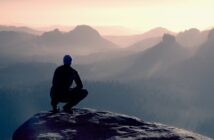 A person wearing a dark jacket and hat squats on the edge of a rocky cliff, overlooking a vast landscape of misty mountains and valleys during sunrise or sunset. The scene has a serene and contemplative atmosphere.