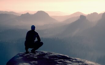 A person wearing a dark jacket and hat squats on the edge of a rocky cliff, overlooking a vast landscape of misty mountains and valleys during sunrise or sunset. The scene has a serene and contemplative atmosphere.