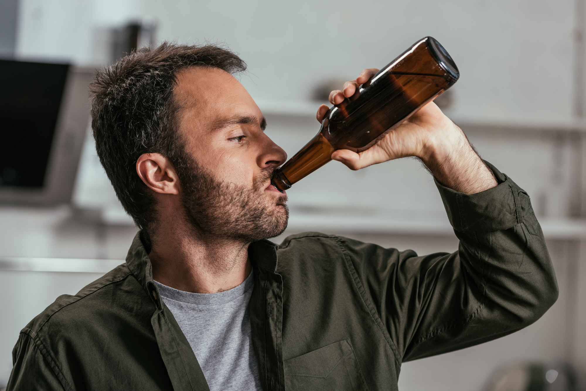 A man with short dark hair and a beard, dressed in a green shirt over a gray t-shirt, is drinking from a brown glass bottle. He is indoors, and the background is blurry.